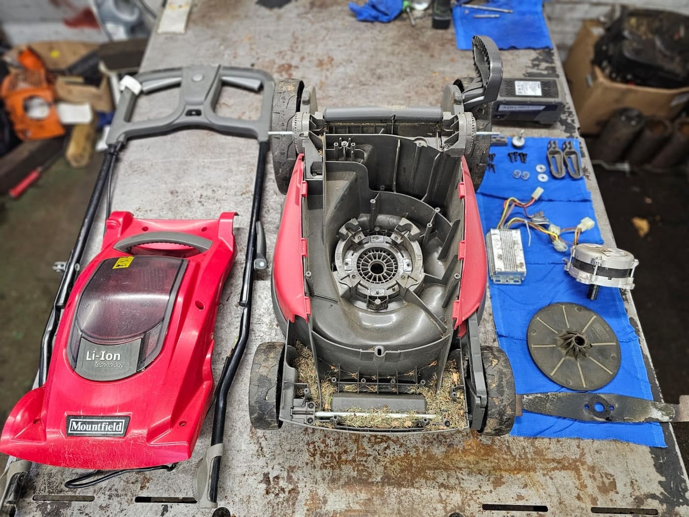 A Mountfield cordless mower stripped down with its parts laid out on the bench